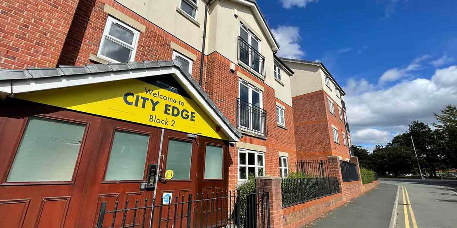 Exterior of City Edge Block 2, a red-brick residential building with a yellow welcome sign, under a partly cloudy sky.