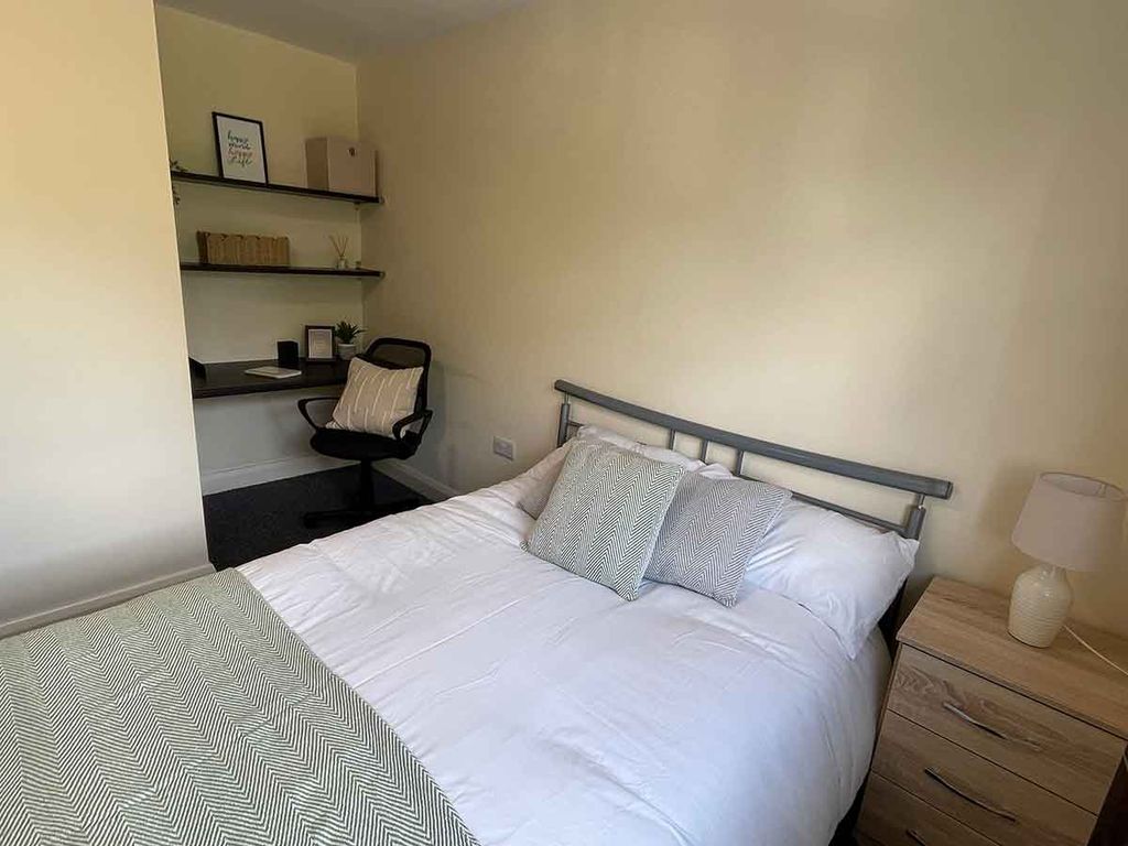 A cozy bedroom with a neatly made bed, two pillows, a bedside table with a lamp, and a small desk with a chair and shelves in the corner.