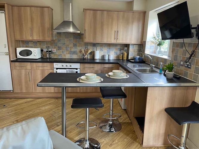 Modern kitchen with wooden cabinets, black countertops, and bar stools. Includes appliances, tiled backsplash, and a wall-mounted TV.