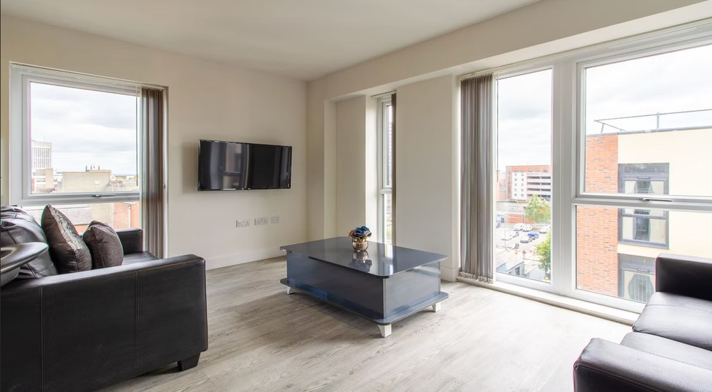 Modern living room with a sleek black sofa, wall-mounted TV, glass coffee table, and large windows offering a city view.