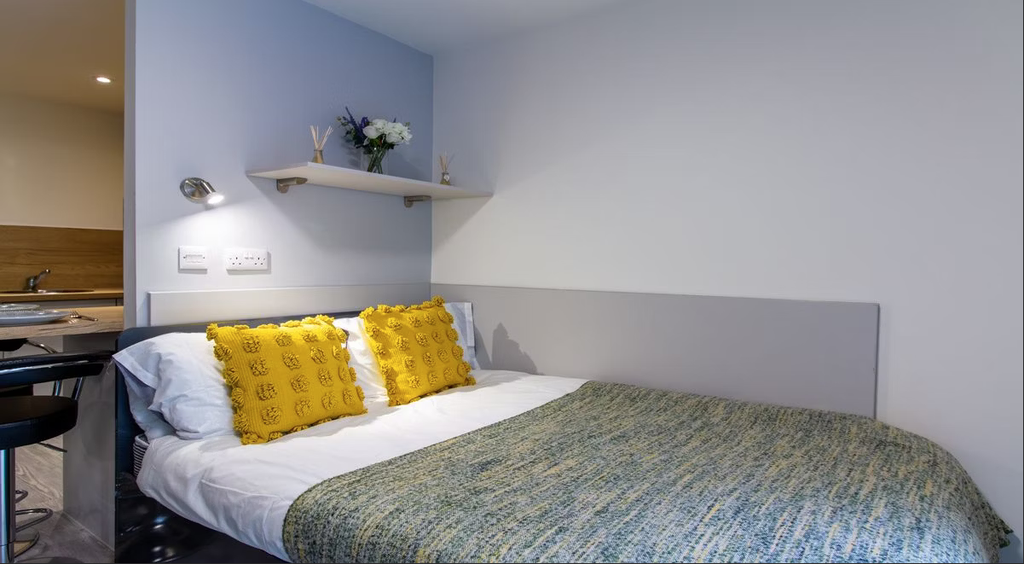 Cozy bedroom with a neatly made bed, yellow pillows, a green throw, wall shelf with decor, and adjacent kitchenette.