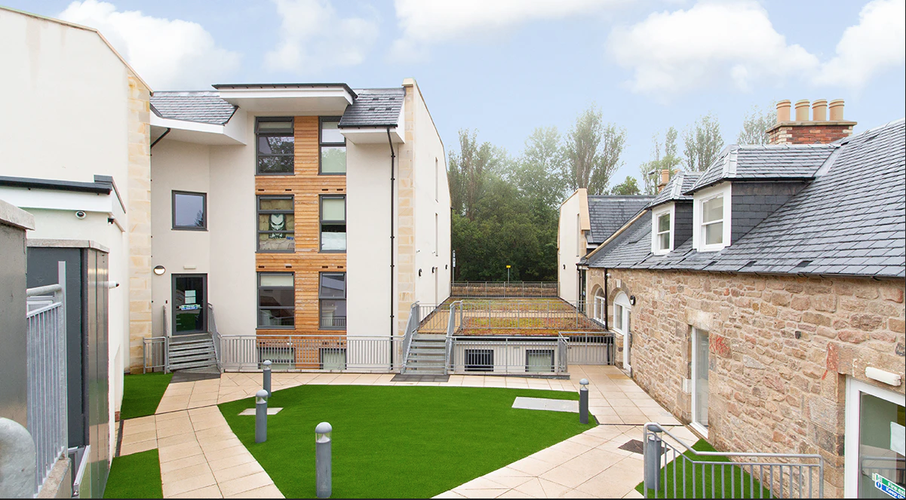 Modern courtyard with a mix of contemporary and traditional buildings, featuring green lawns, pathways, and a backdrop of trees.