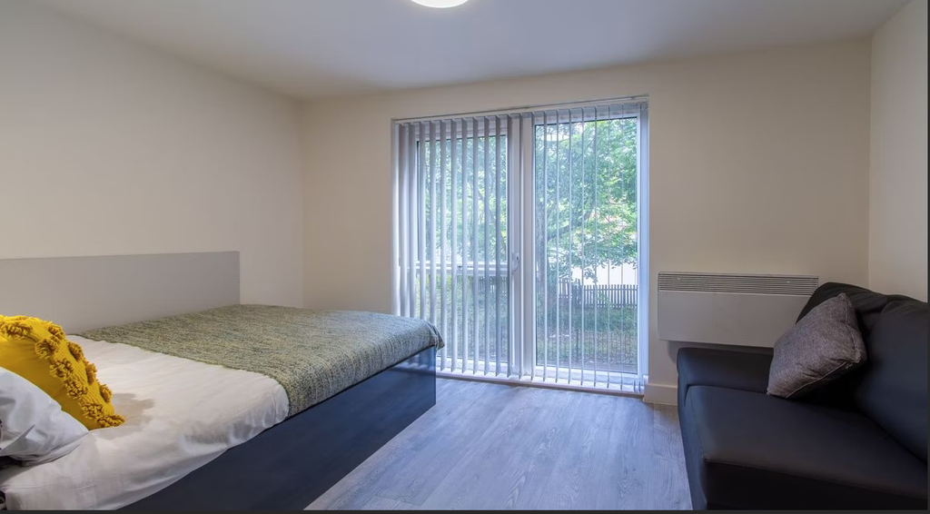 Minimalist bedroom with a bed, green throw, yellow pillow, sofa, and large window with vertical blinds overlooking greenery.
