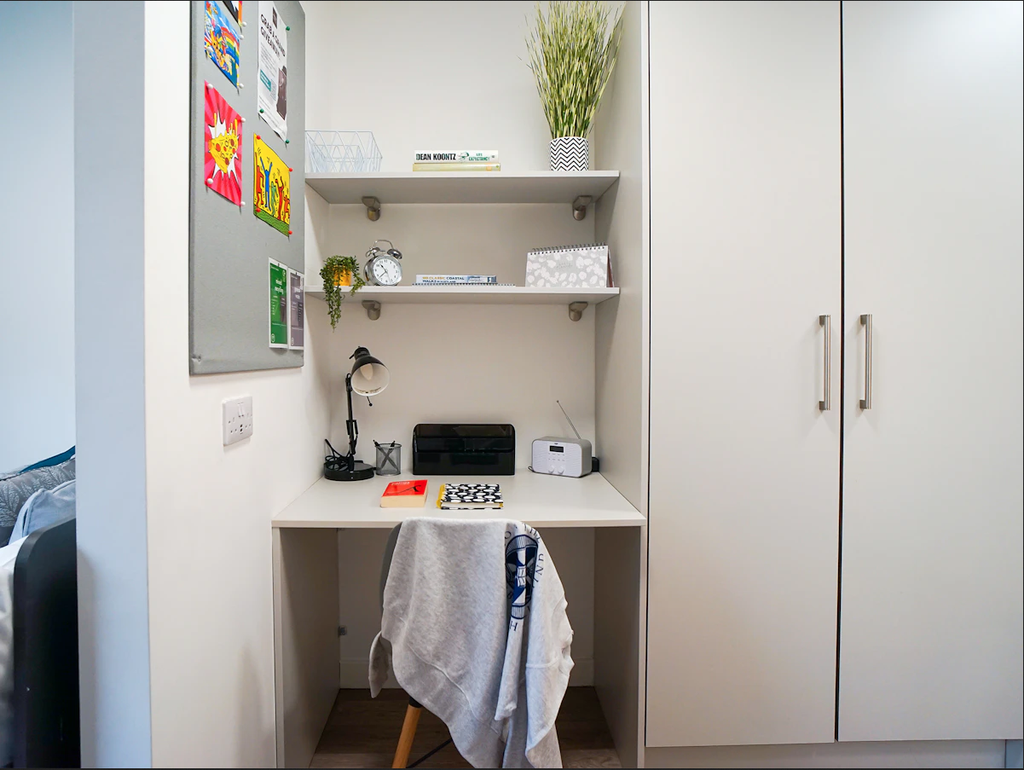 Compact workspace with a desk, chair, and shelves. Items include a lamp, printer, clock, books, and decorative plants. A bulletin board is on the left wall.