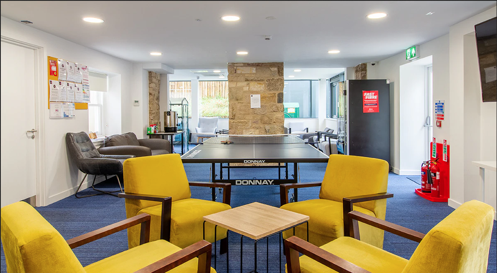 Modern lounge with a ping pong table, yellow chairs around a small table, and a cozy seating area. Bright lighting and stone wall accents.