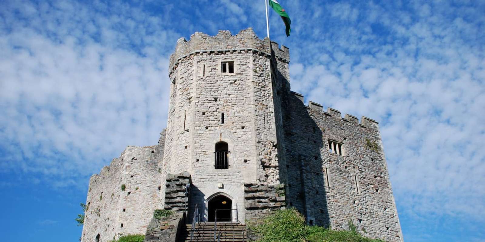 Cardiff castle tower with a green flag on top, set against a blue sky with scattered clouds, viewed from below with steps leading up.