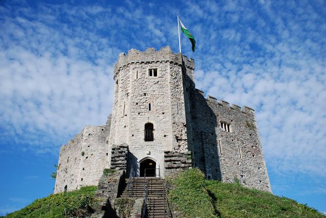 Cardiff castle tower with a green flag on top, set against a blue sky with scattered clouds, viewed from below with steps leading up.