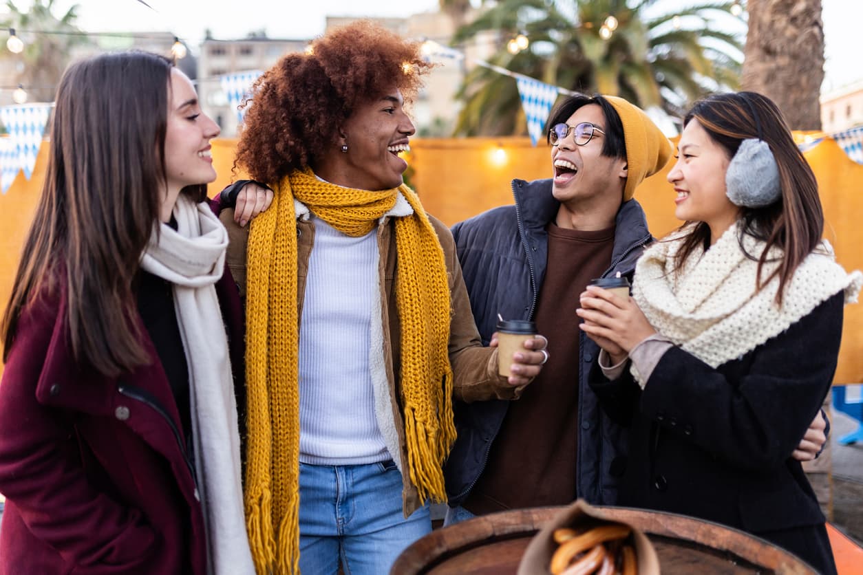 Four friends in winter clothing laugh and chat, holding hot drinks at an outdoor market with festive lights and palm trees in the background.
