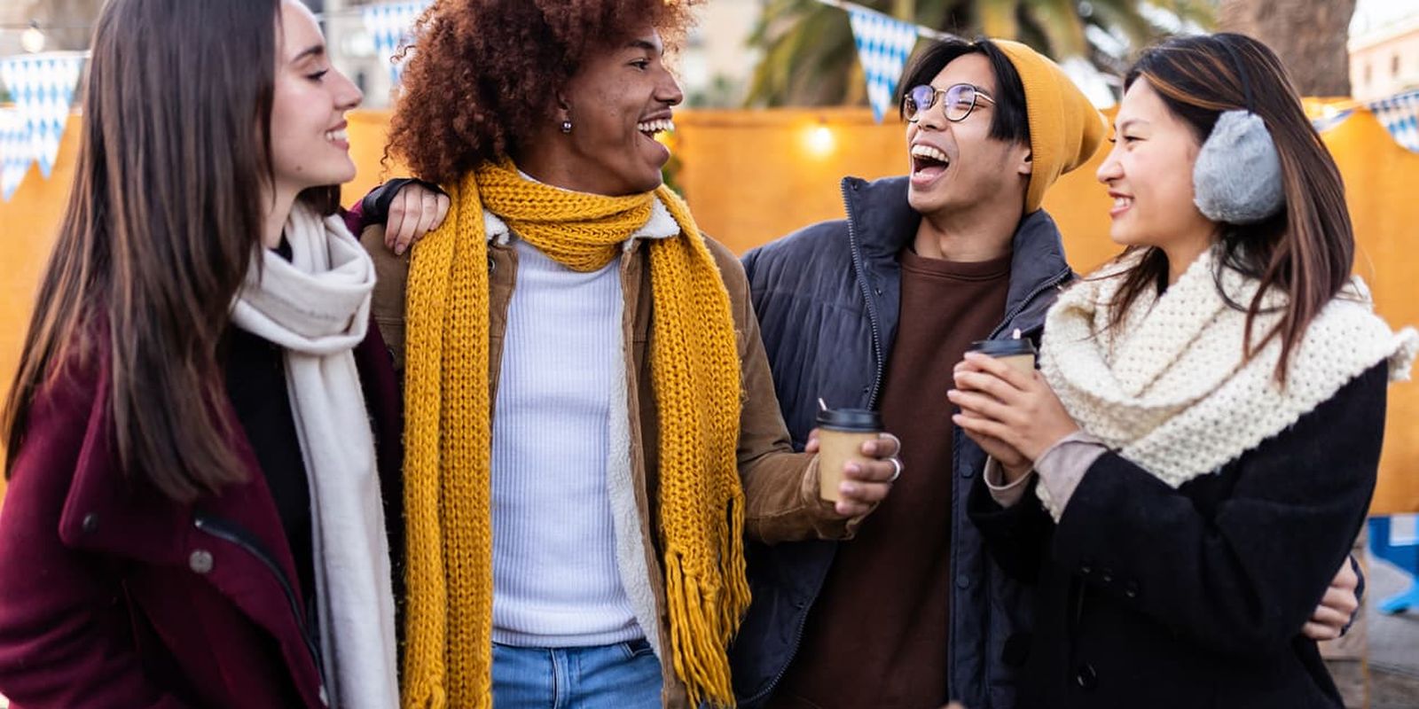 Four friends in winter clothing laugh and chat, holding hot drinks at an outdoor market with festive lights and palm trees in the background.