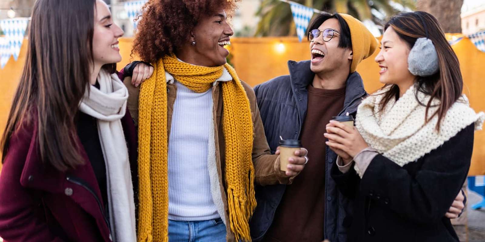 Four friends in winter clothing laugh and chat, holding hot drinks at an outdoor market with festive lights and palm trees in the background.