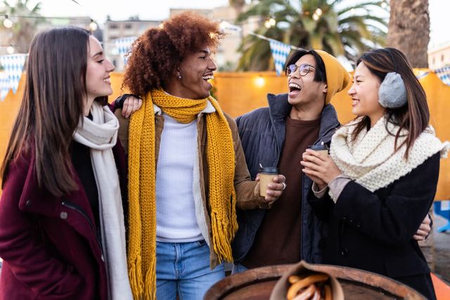 Four friends in winter clothing laugh and chat, holding hot drinks at an outdoor market with festive lights and palm trees in the background.