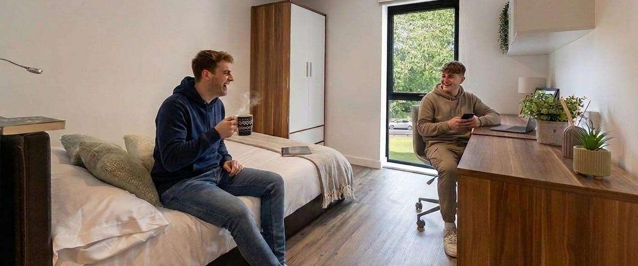 Two people in a cozy room, one sitting on a bed with a mug, and the other at a desk with a phone, both smiling, near a large window.