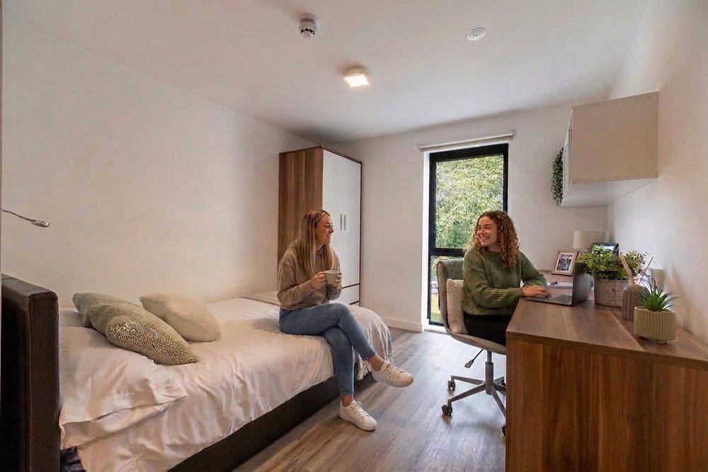 Two women in a tidy dorm room; one sits on a bed holding a mug, and the other sits at a desk with a laptop, both smiling.