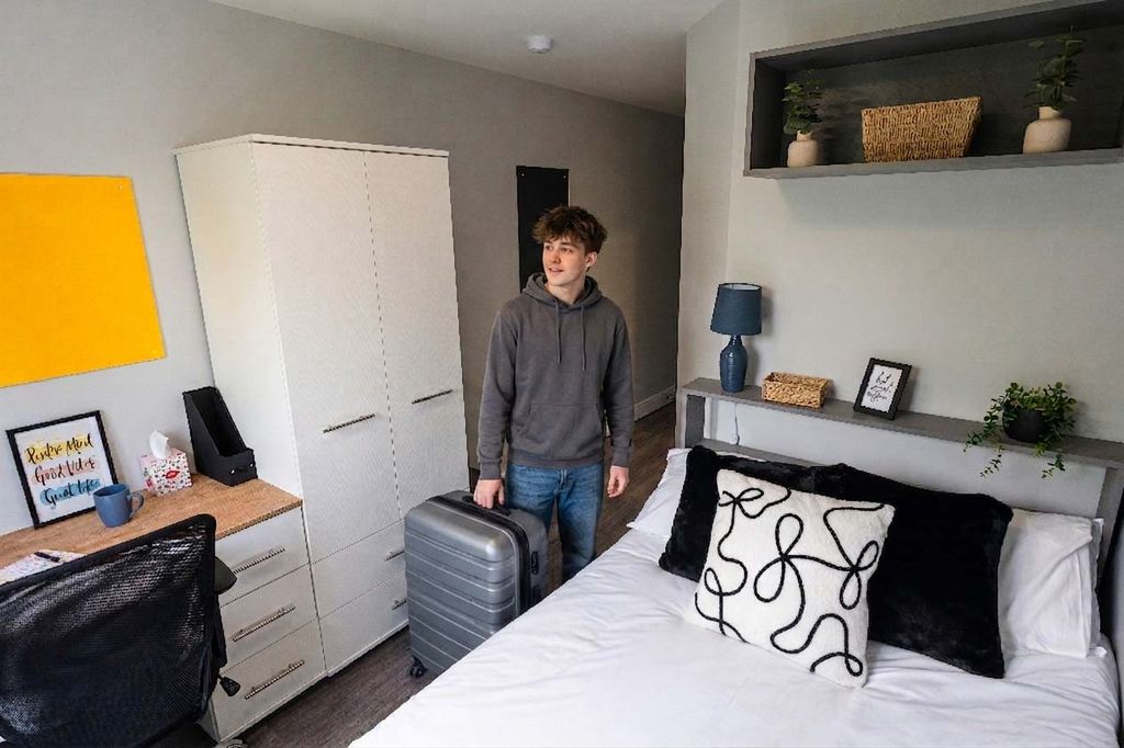 A person stands in a modern bedroom with a suitcase, featuring a bed, desk, and decorative shelves.