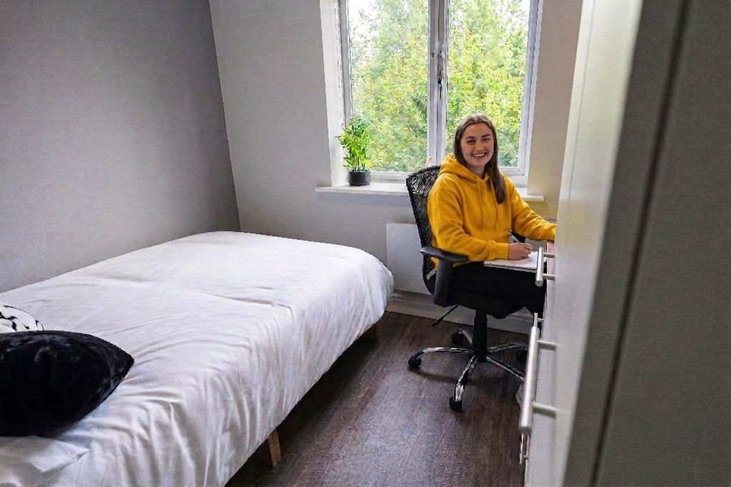 A person in a yellow hoodie sits at a desk in a small, bright bedroom with a bed and a window with greenery outside.