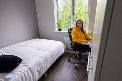 A person in a yellow hoodie sits at a desk in a small, bright bedroom with a bed and a window with greenery outside.