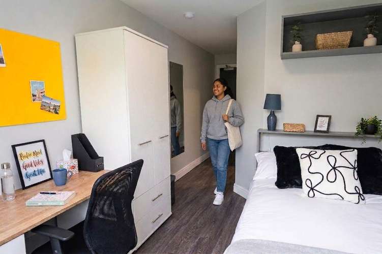 A person in a hoodie and jeans walks into a tidy, modern room featuring a bed, desk, chair, and decorative items on shelves and walls.
