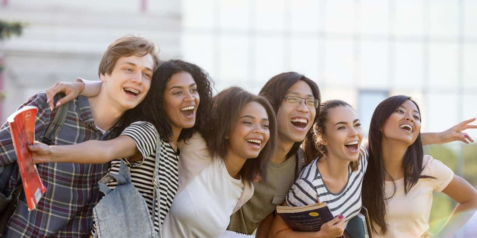 A diverse group of six joyful students taking a selfie outdoors, holding books and backpacks, with expressions of laughter and excitement.