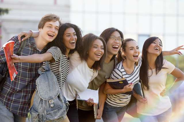 A diverse group of six joyful students taking a selfie outdoors, holding books and backpacks, with expressions of laughter and excitement.