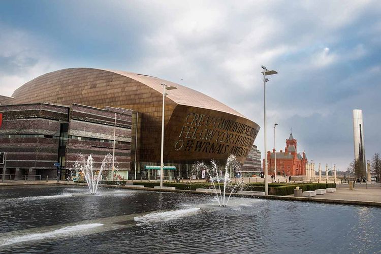 The image shows the Wales Millennium Centre with its distinctive bronze dome and large lettering, set against a cloudy sky, with fountains in front.