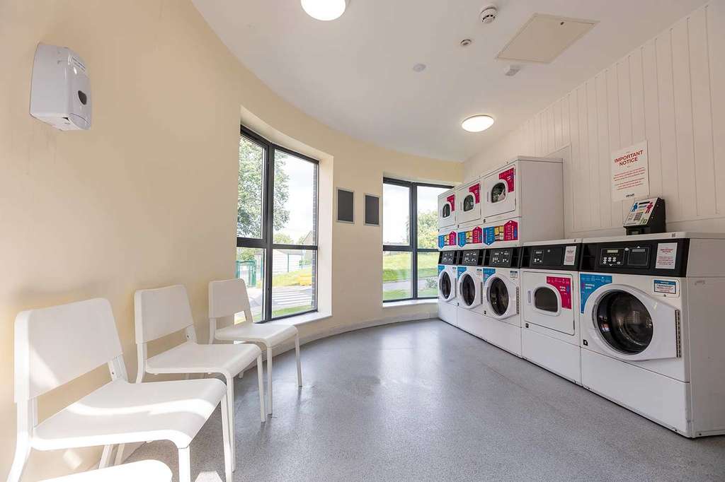 Bright laundry room with large windows, chairs, and a row of washing machines and dryers. White walls and a light floor create an airy atmosphere.
