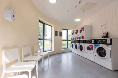 Bright laundry room with large windows, chairs, and a row of washing machines and dryers. White walls and a light floor create an airy atmosphere.
