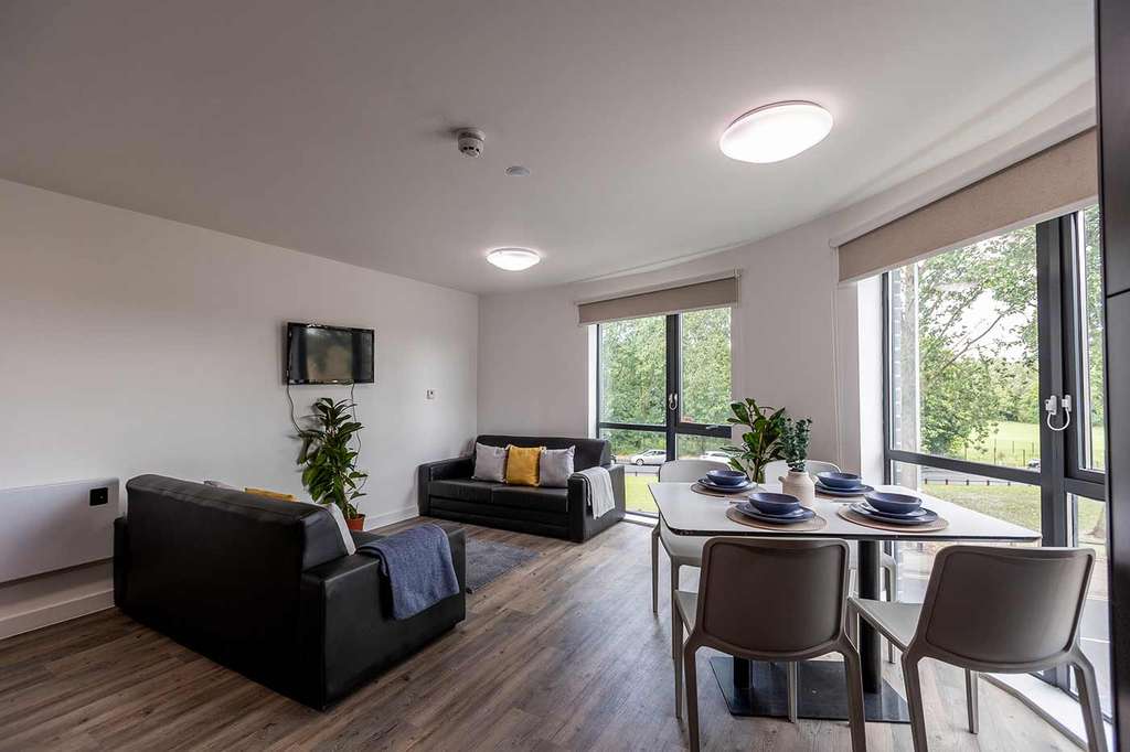 Modern living room with two sofas, a wall-mounted TV, dining table set for four, and large windows overlooking greenery.