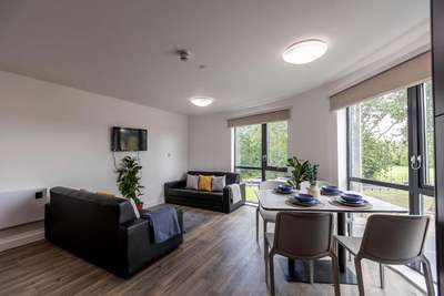 Modern living room with two sofas, a wall-mounted TV, dining table set for four, and large windows overlooking greenery.