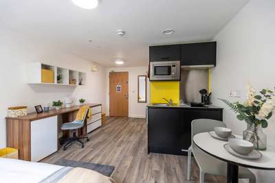 Modern studio apartment with a kitchenette, dining table, desk, and bed. Features wood flooring, white walls, and decorative shelves.