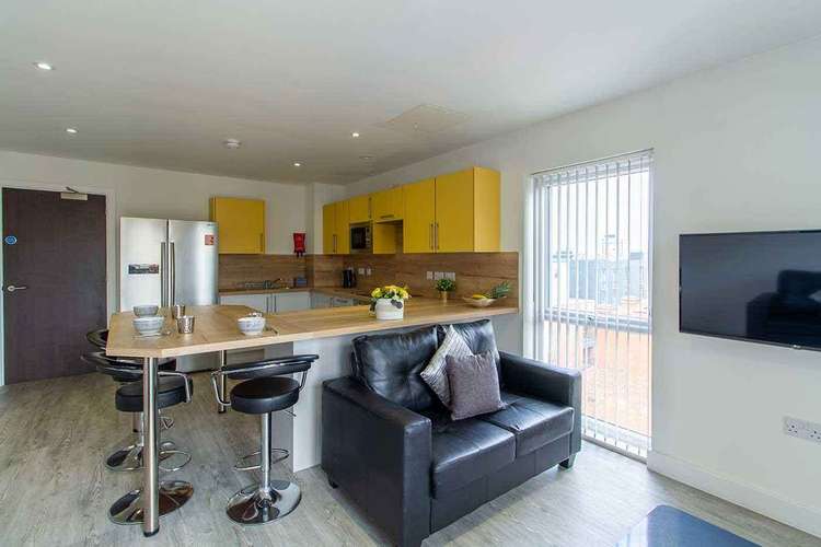 Modern kitchen and living area with yellow cabinets, stainless steel appliances, a black sofa, bar stools, and a wall-mounted TV.