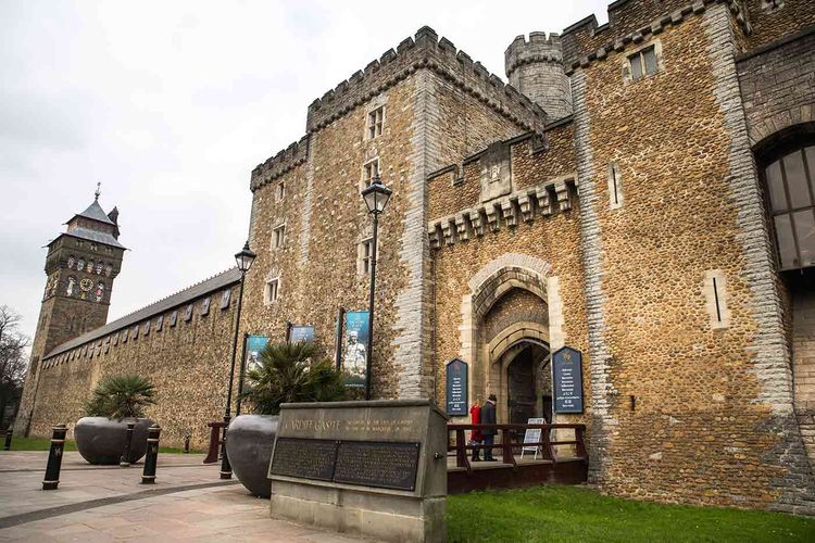 Historic stone castle with a clock tower, arched entrance, and informational signs. Pathway and large planters in the foreground.