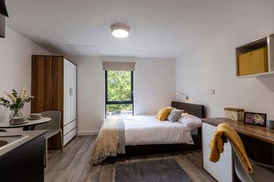 Modern bedroom with a bed, wooden desk, wardrobe, and window overlooking greenery. Decor includes a yellow box and a vase with flowers.