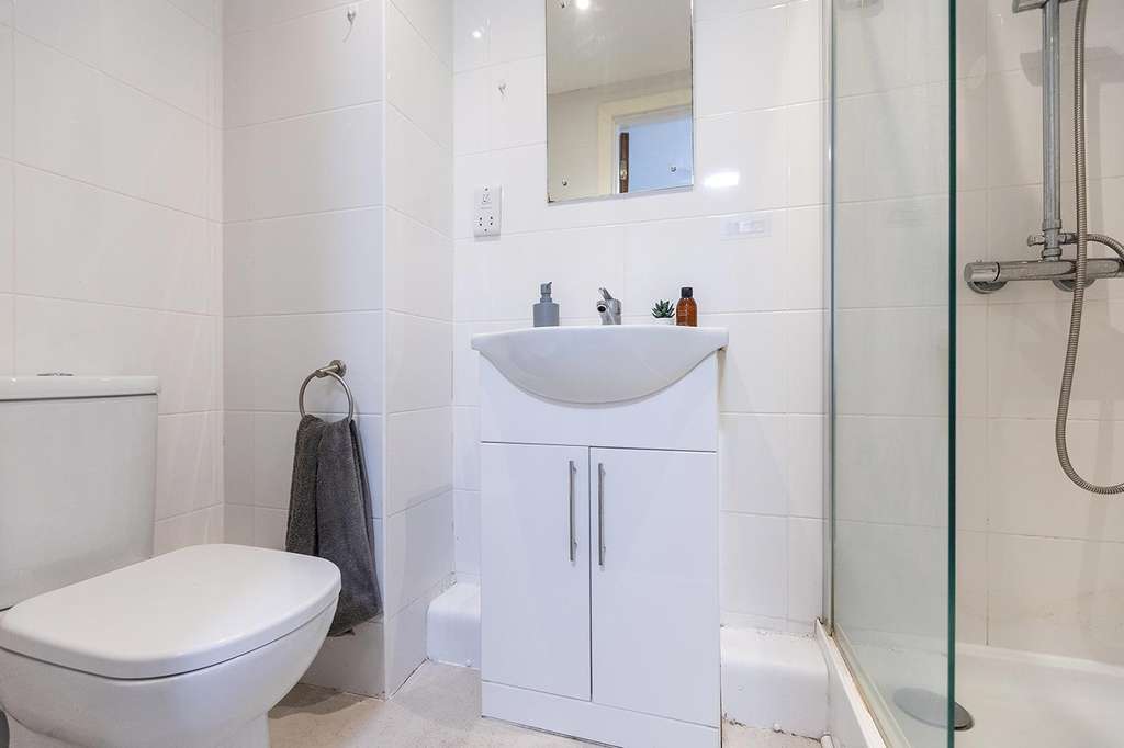 Modern bathroom with white tiles, featuring a toilet, sink with cabinet, mirror, towel rack, and glass-enclosed shower.