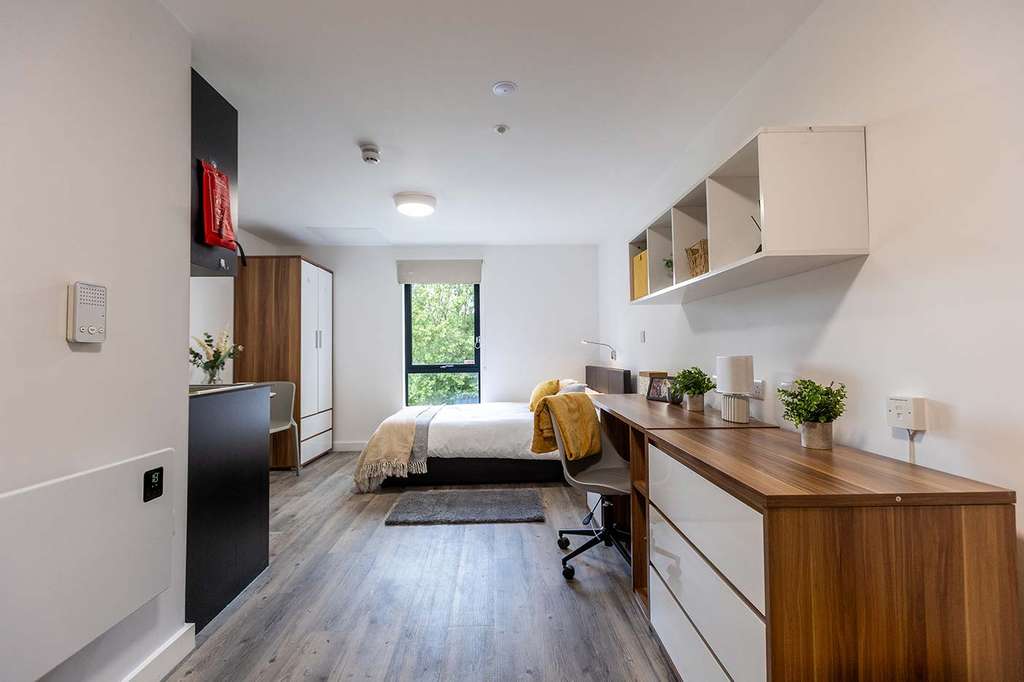 Modern, minimalist bedroom with a bed, wooden desk, chair, shelves, and a window overlooking greenery. Light walls and wood flooring.