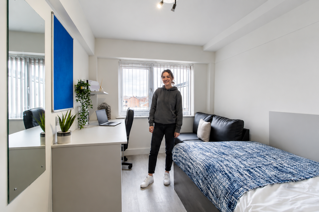 A person stands in a bright, modern bedroom with a bed, desk, laptop, and plants. The room has a large window and a blue bulletin board.