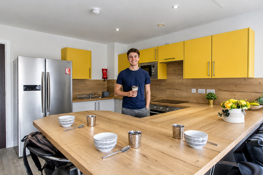 A man holding a takeout cup smiles in a modern kitchen with yellow cabinets, a large wooden counter, and place settings on the table.