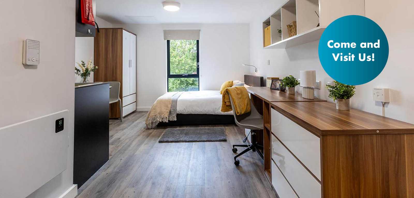 Modern studio apartment with a bed, kitchenette, dining area, wardrobe, and a window overlooking greenery. Sign reads "Come and Visit Us!"