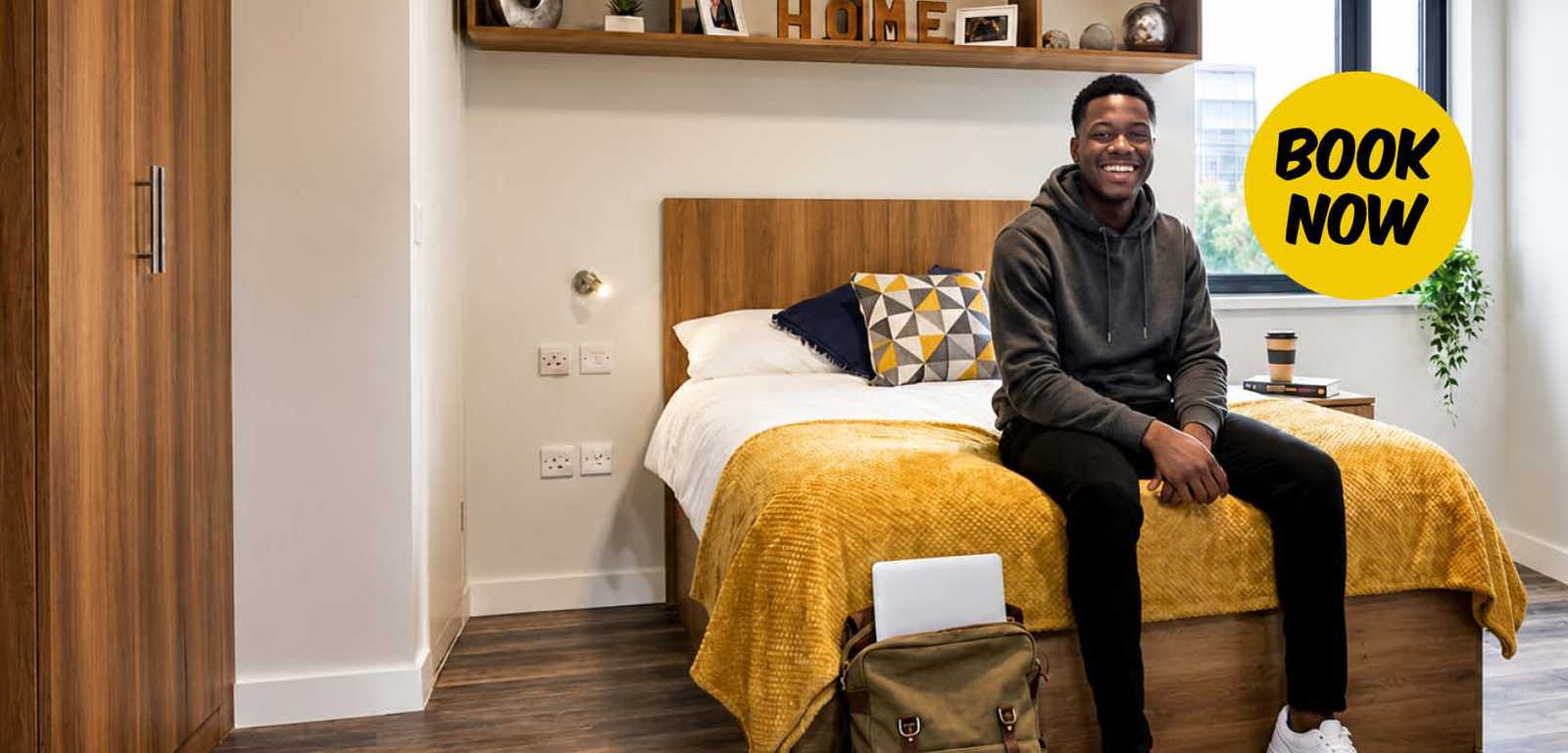 A person sitting on a bed in a modern, tidy room, with a yellow "Book Now" sign. A backpack and laptop are on the floor, a coffee cup nearby.