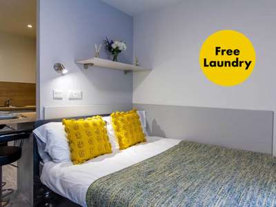 A cozy bedroom with a neatly made bed, two yellow pillows, bedside lighting, a shelf with flowers, and a sign indicating free laundry.