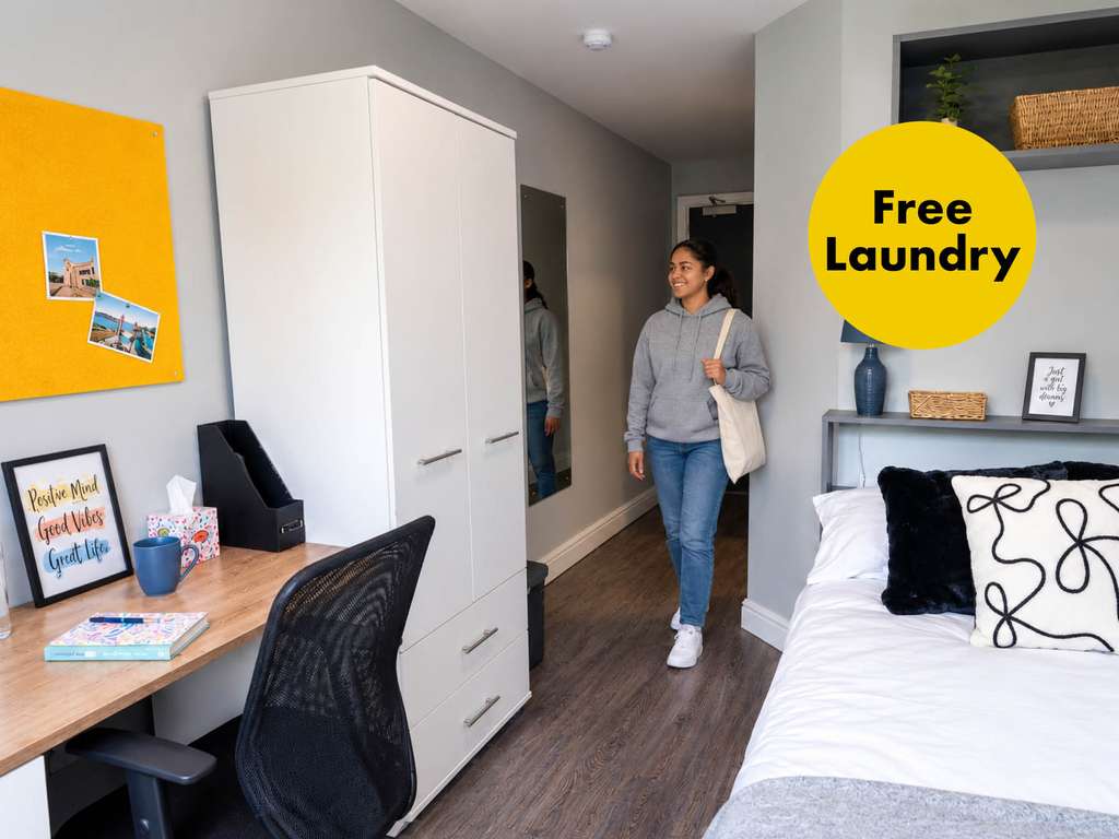 A person in a gray hoodie walks into a tidy bedroom, featuring a bed, a desk with a chair, and a "Free Laundry" sign on the wall.