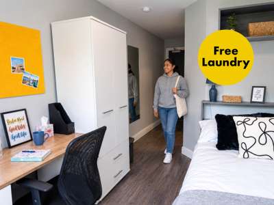 A person in a gray hoodie walks into a tidy bedroom, featuring a bed, a desk with a chair, and a "Free Laundry" sign on the wall.