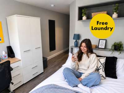A woman sits on a bed in a cozy, modern room, smiling at her phone. A large yellow circle reads "Free Laundry."