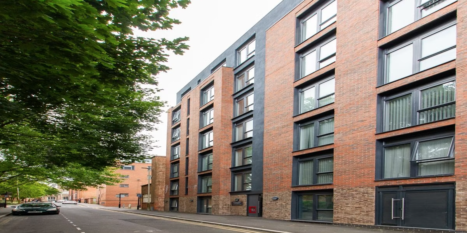 A modern, multi-story brick apartment building with large windows on a tree-lined street.