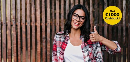 Smiling woman in glasses and plaid shirt gives thumbs up; yellow circle with "Get £1000 Cashback!" text on wooden background.
