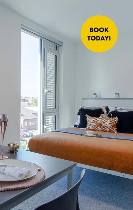 Bright bedroom with a large window, orange bedspread, decorative pillows, and a small table set for dining. Yellow "Book Today!" circle in corner.