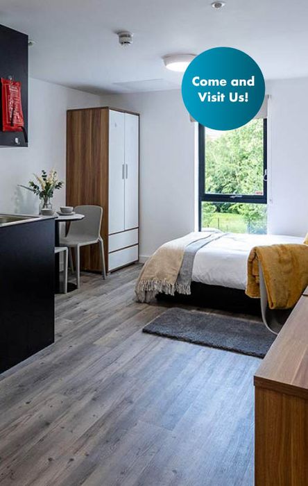 Modern studio apartment with a bed, kitchenette, dining area, wardrobe, and a window overlooking greenery. Sign reads "Come and Visit Us!"