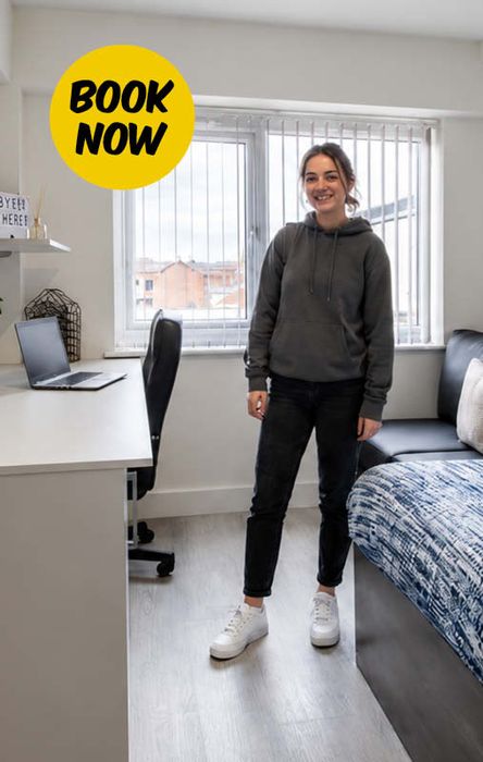 Person in a hoodie stands smiling in a tidy, modern bedroom with a desk, chair, and laptop. A "Book Now" icon is displayed above.