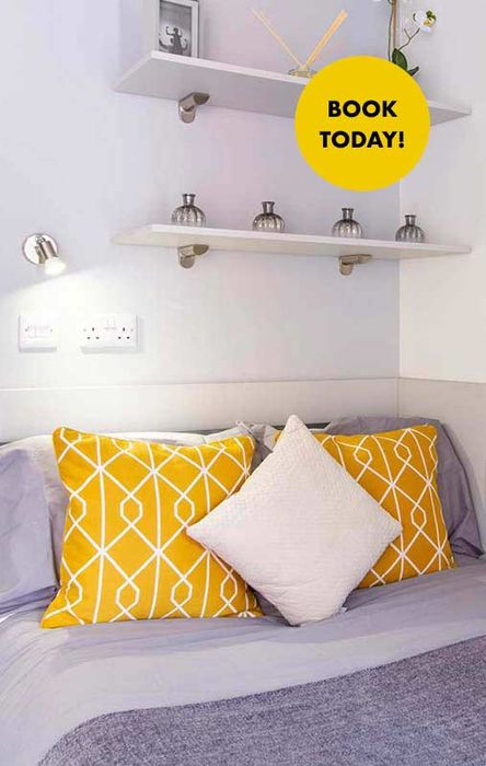 Cozy bedroom with yellow and white pillows, wall-mounted shelves, small decor items, and a "Book Today!" sign.