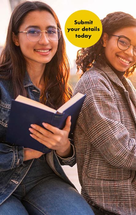 Two people sitting outdoors, one holding a book. Both are smiling and wearing glasses. A yellow circle reads, "Submit your details today."