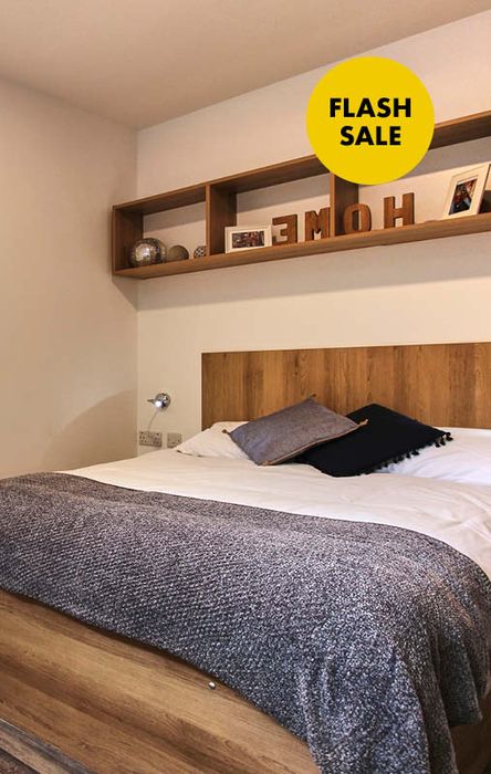 Cozy bedroom with a wooden bed, gray and white bedding, decorative pillows, and a shelf with decor. A "Flash Sale" sign is in the corner.
